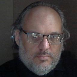 A smiling grey-bearded man wearing glasses and a black shirt with a backdrop fo a corner of a darkened room and doors with light from a casement window shining through space between those doors.